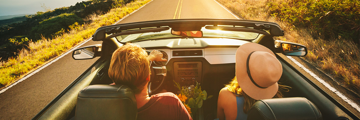 Two people ride in a convertible along a sunlit rural road at golden hour, viewed from behind, with one passenger wearing a wide-brimmed hat and flowers between the seats
