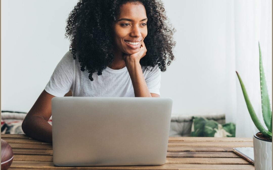 A smiling woman with curly hair rests her chin on her hand while using a laptop at a wooden table next to a potted aloe plant