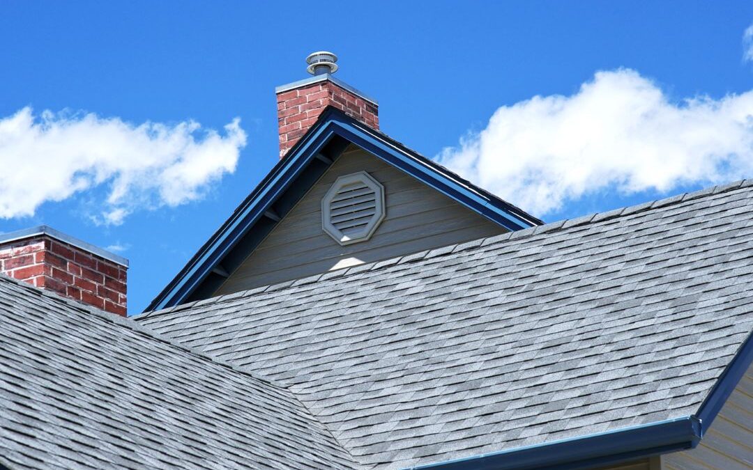 Low-angle upward view of a house's grey asphalt shingled roof featuring red brick chimneys, an octagonal gable vent, and a bright blue sky dotted with fluffy white clouds