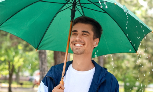 A smiling young man stands outdoors under a large green umbrella as rain falls - www.lisafrenchinsurance.com
